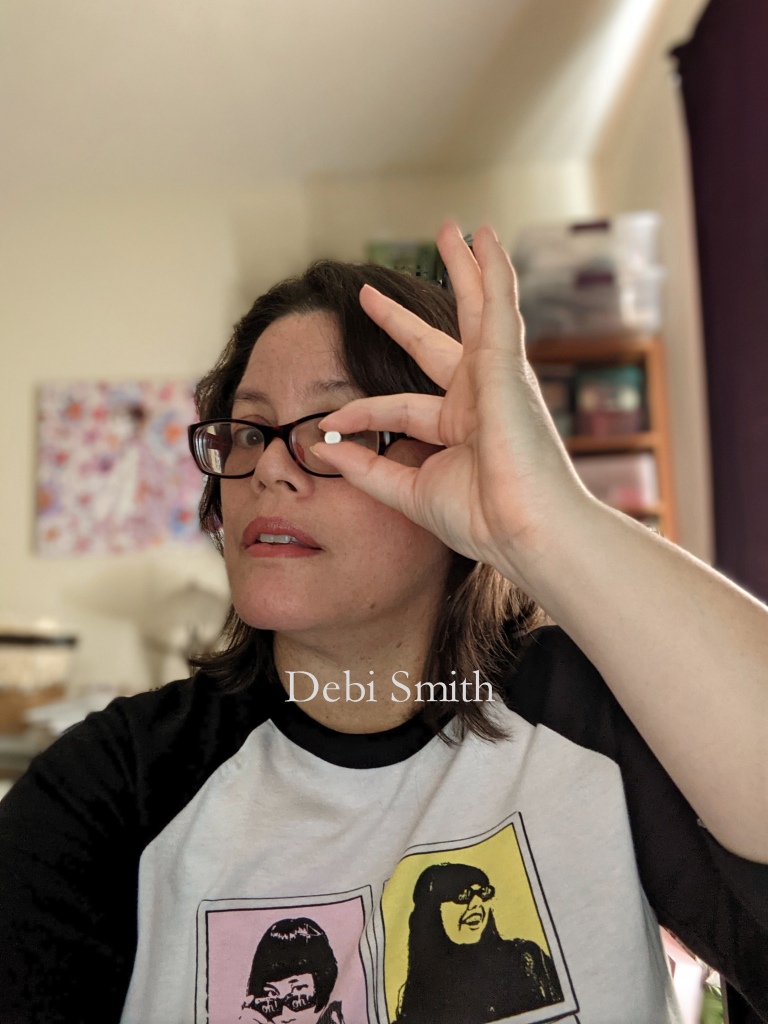 Photo of author holding up a tiny pill of Adderall between her thumb an forefinger in front of her left eye. She is wearing black-rimmed, rectangular glasses and a baseball style shirt with black sleeves and artistic images of each band member of The Linda Lindas in 4 square panels. The author has medium length dark brown hair parted in the middle, brown eyes, and tan skin with freckles.